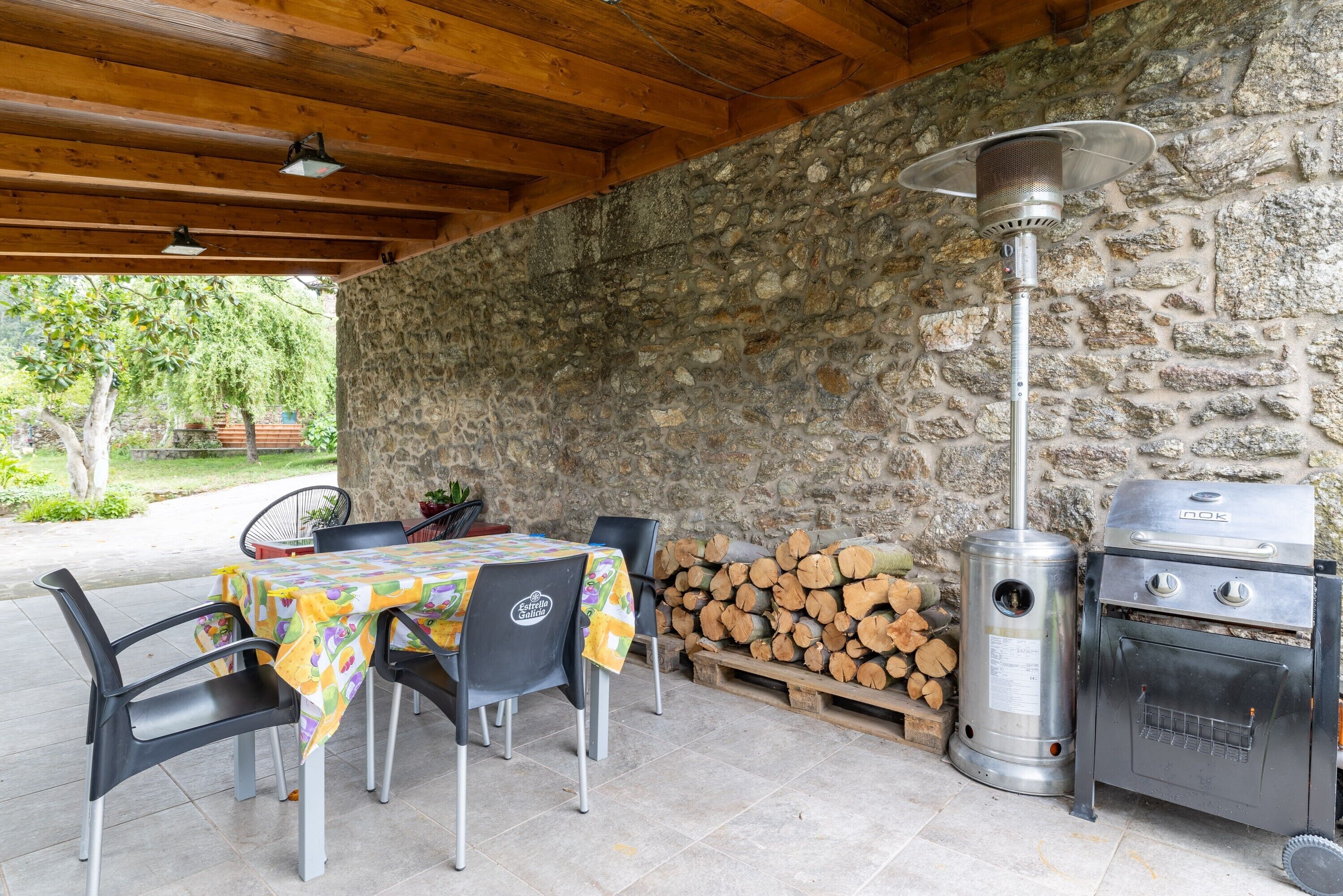 Een terras buiten met een stenen muur, een barbecue, een tafel met stoelen en een terrasverwarmer.