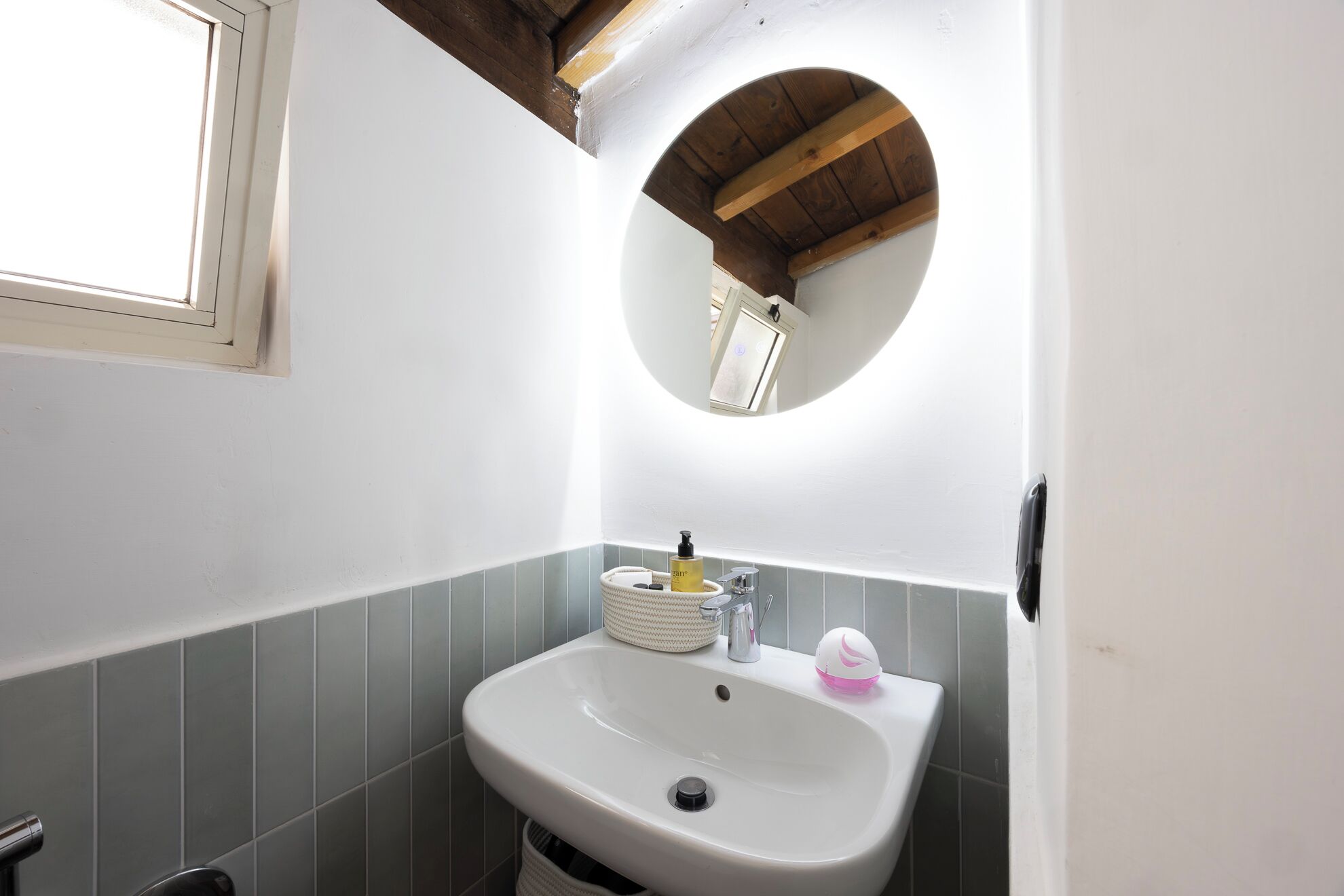 A bathroom with a white sink, a round mirror, and a window.