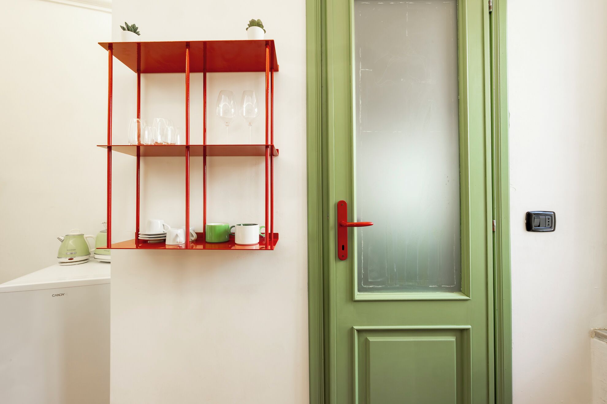 Una puerta verde con un panel de vidrio esmerilado, un picaporte rojo y un estante rojo con cristalería y vajilla.