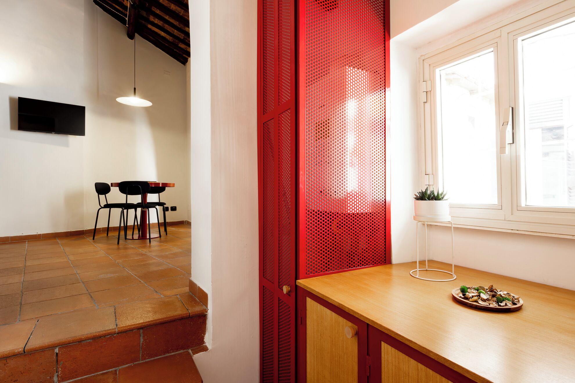 A room with a wooden table, a window with a plant, a red mesh partition, and a staircase.
