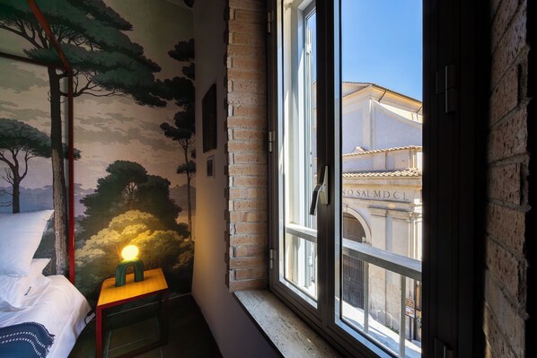 A room with a bed, a small table with a lamp, a mural of trees, and a window with a view of a building.