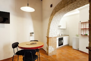 A dining area with a round table, chairs, a pendant light, and a stone archway leading to a kitchen.