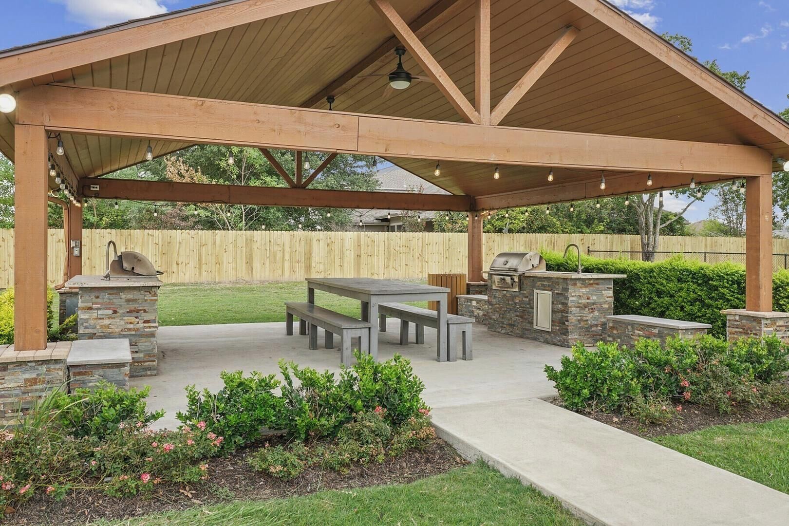Um pátio ao ar livre com churrasqueira, estação de churrasco em pedra e pérgola de madeira com luzes.