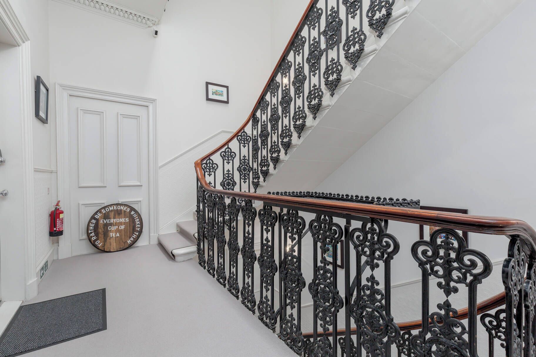 Un escalier orné de rampes en métal noir et d’une main courante en bois, menant à une porte blanche surmontée d’un tableau encadré.