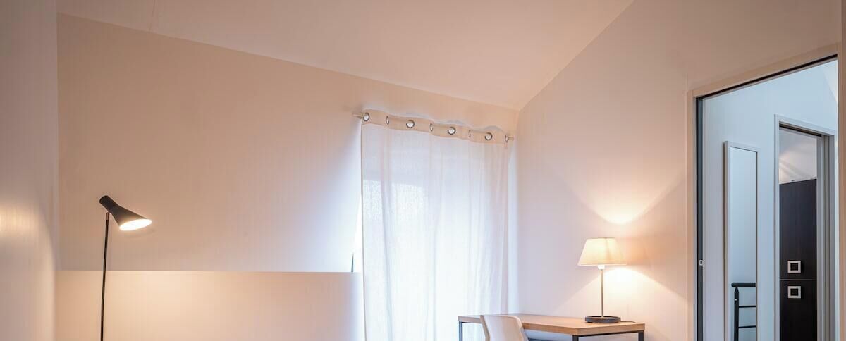 A hotel room with a bed, a desk, a chair, a lamp, and a window with curtains.