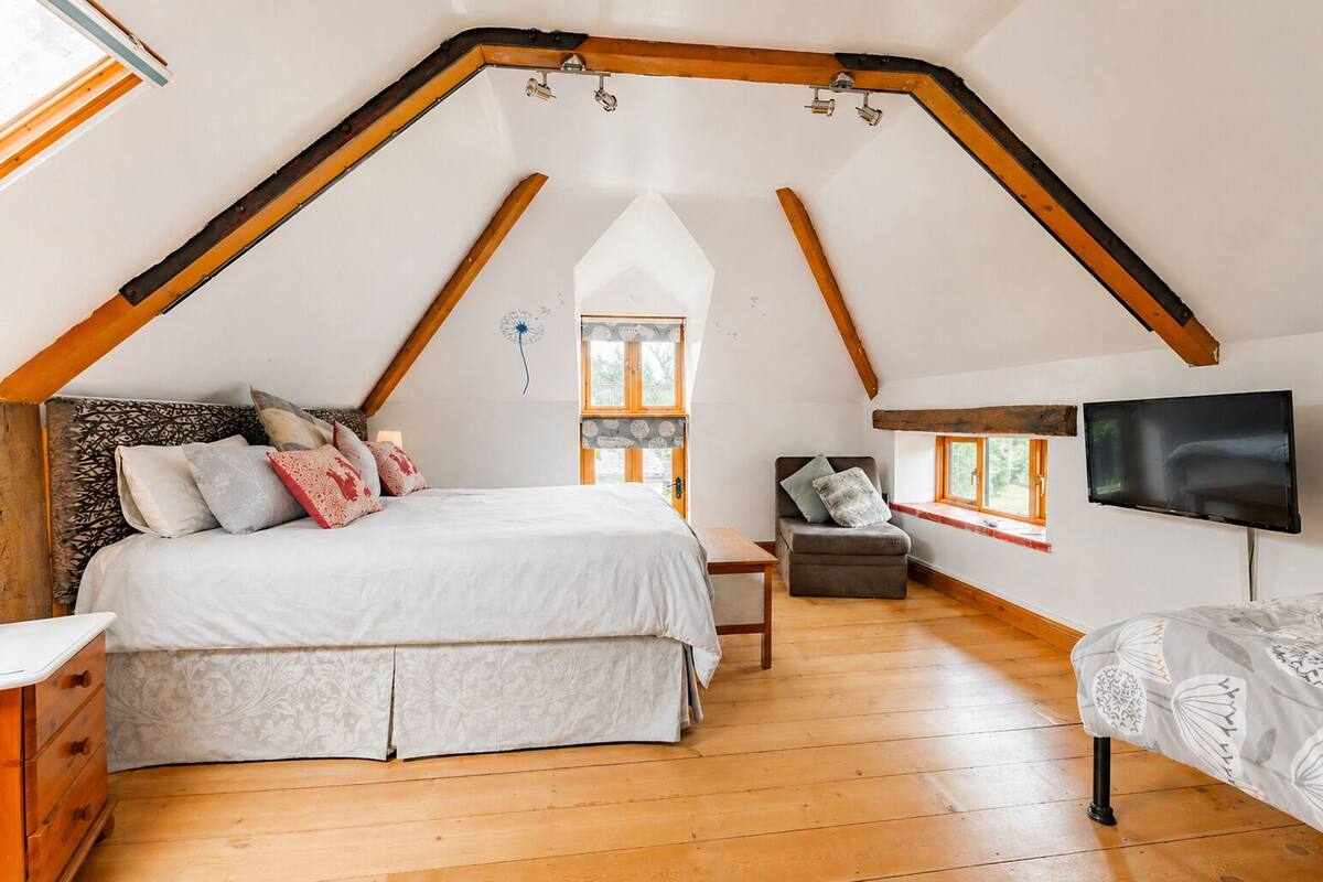 Un dormitorio con techo inclinado, cama, silla, televisor y ventana.