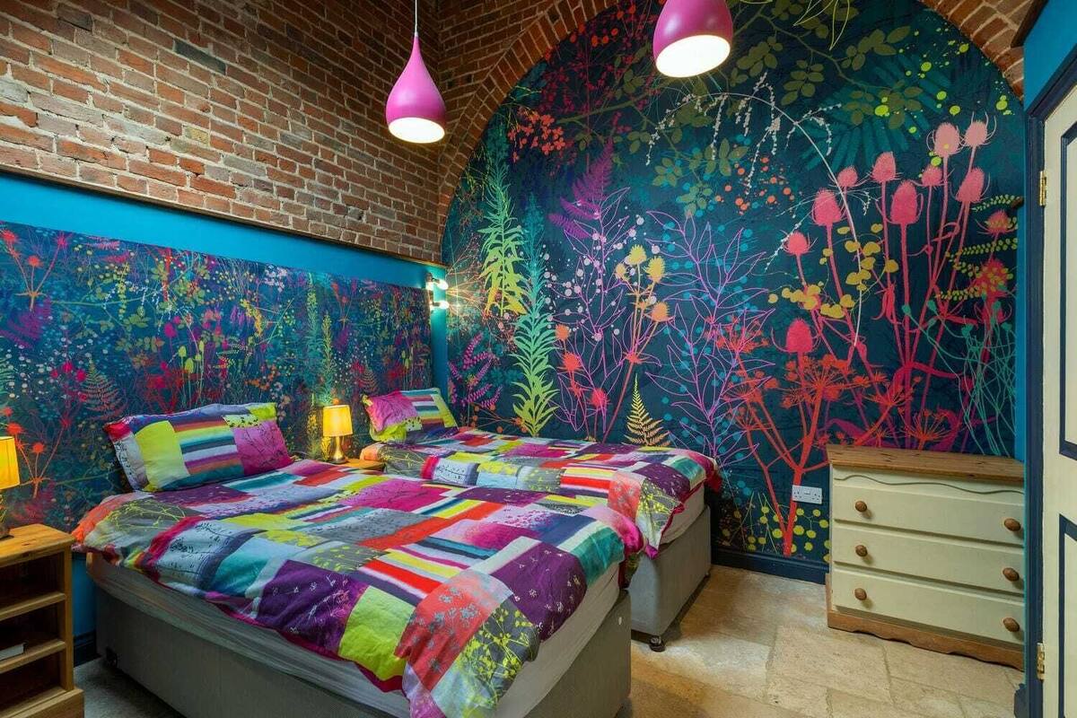 Un dormitorio con pared de ladrillo, un mural colorido, una cama con colcha multicolora y dos lámparas colgantes.