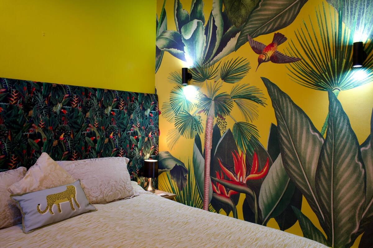 Una habitación con cama, almohadas, mesita de noche y un mural tropical en la pared.