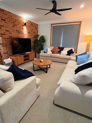 A living room with a brick wall, a flat-screen TV, a ceiling fan, two sofas, a coffee table, and a large window with blinds. - Top Deck (Sawtell)