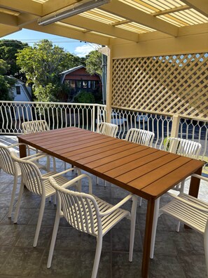 Eine überdachte Terrasse im Freien mit einem Holztisch und weißen Plastikstühlen.