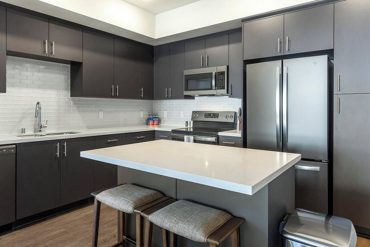 A modern kitchen with dark cabinetry, a white countertop, stainless steel appliances, and two bar stools.