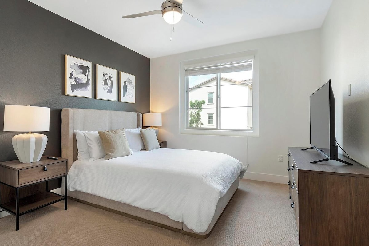 A bedroom with a bed, two bedside lamps, a ceiling fan, a TV on a wooden cabinet, and a window with a view of buildings.