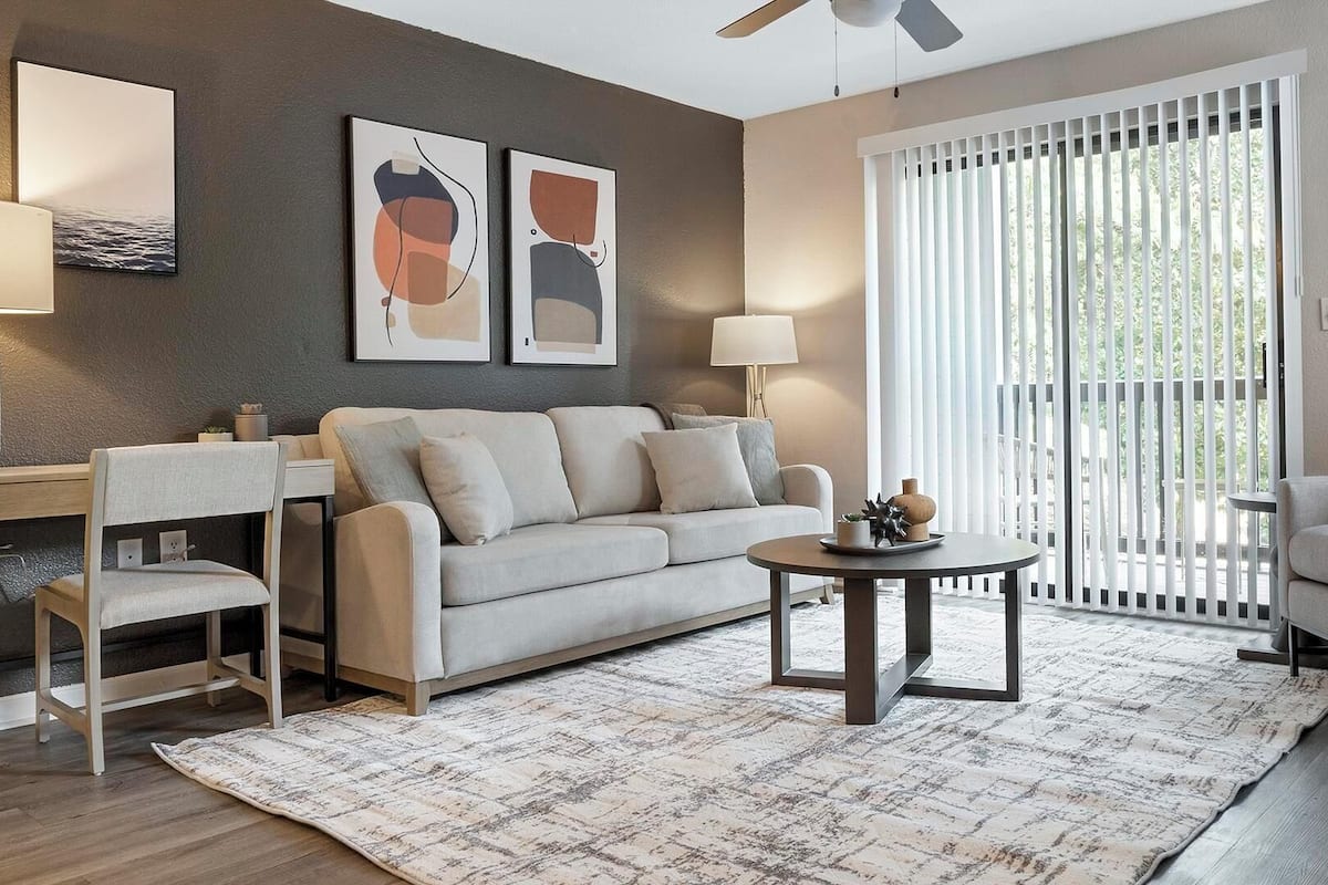A modern living room with a sofa, a coffee table, and a desk. There are two abstract paintings on the wall, a ceiling fan, and a window with blinds.