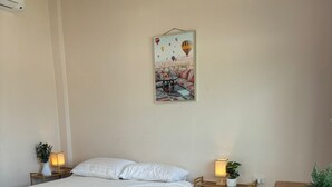 Um quarto com uma cama, mesas de cabeceira, uma pequena mesa com uma cadeira, um ar condicionado fixado na parede e um quadro emoldurado.