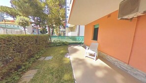 A balcony with a white chair, a hedge, and a building with an orange wall and a window. - Two-story Family Flat With Terrace (Lignano Sabbiadoro)