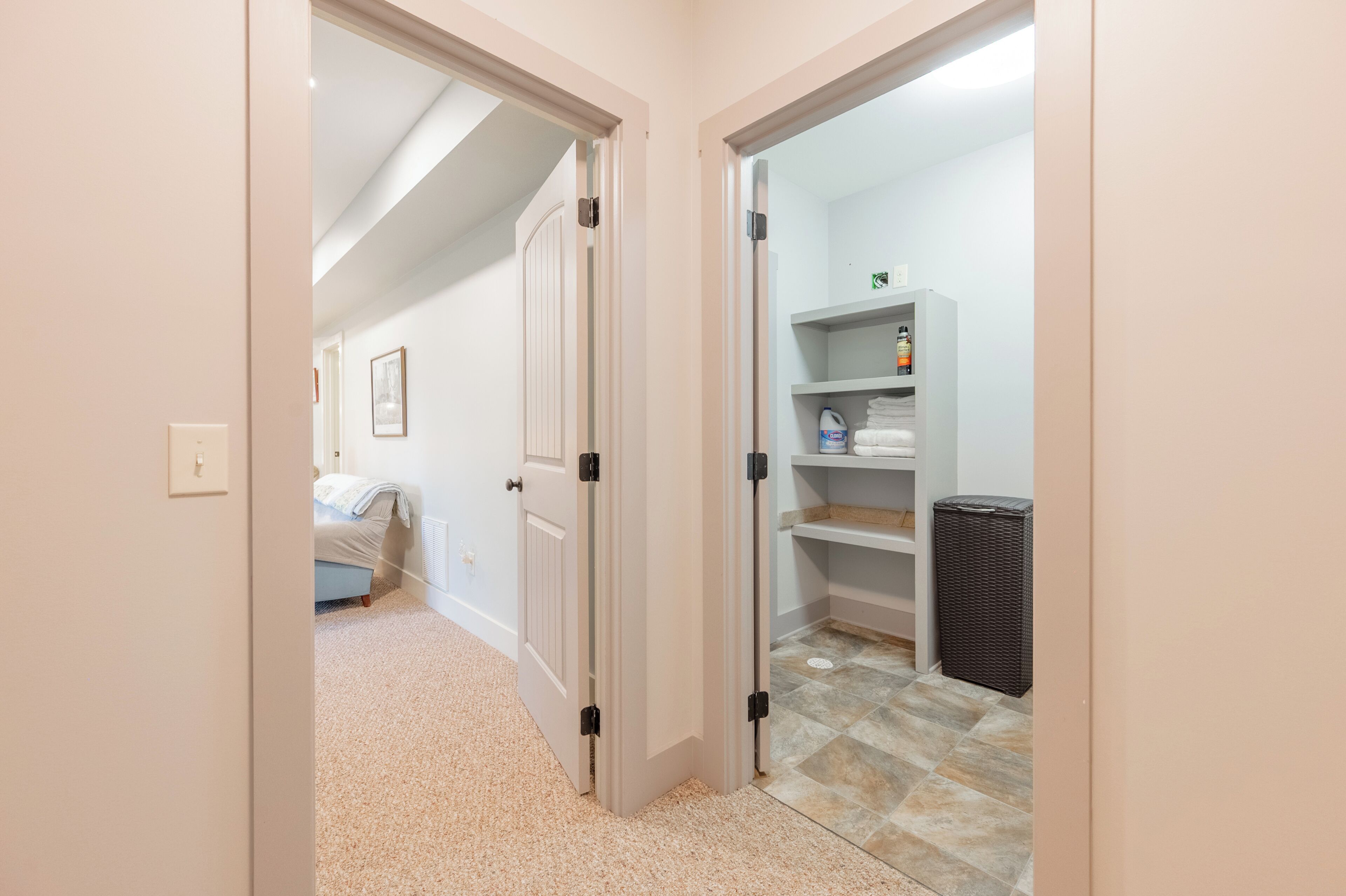 Um corredor com uma cama, uma porta branca e uma área de lavandaria com prateleiras e uma cesta.