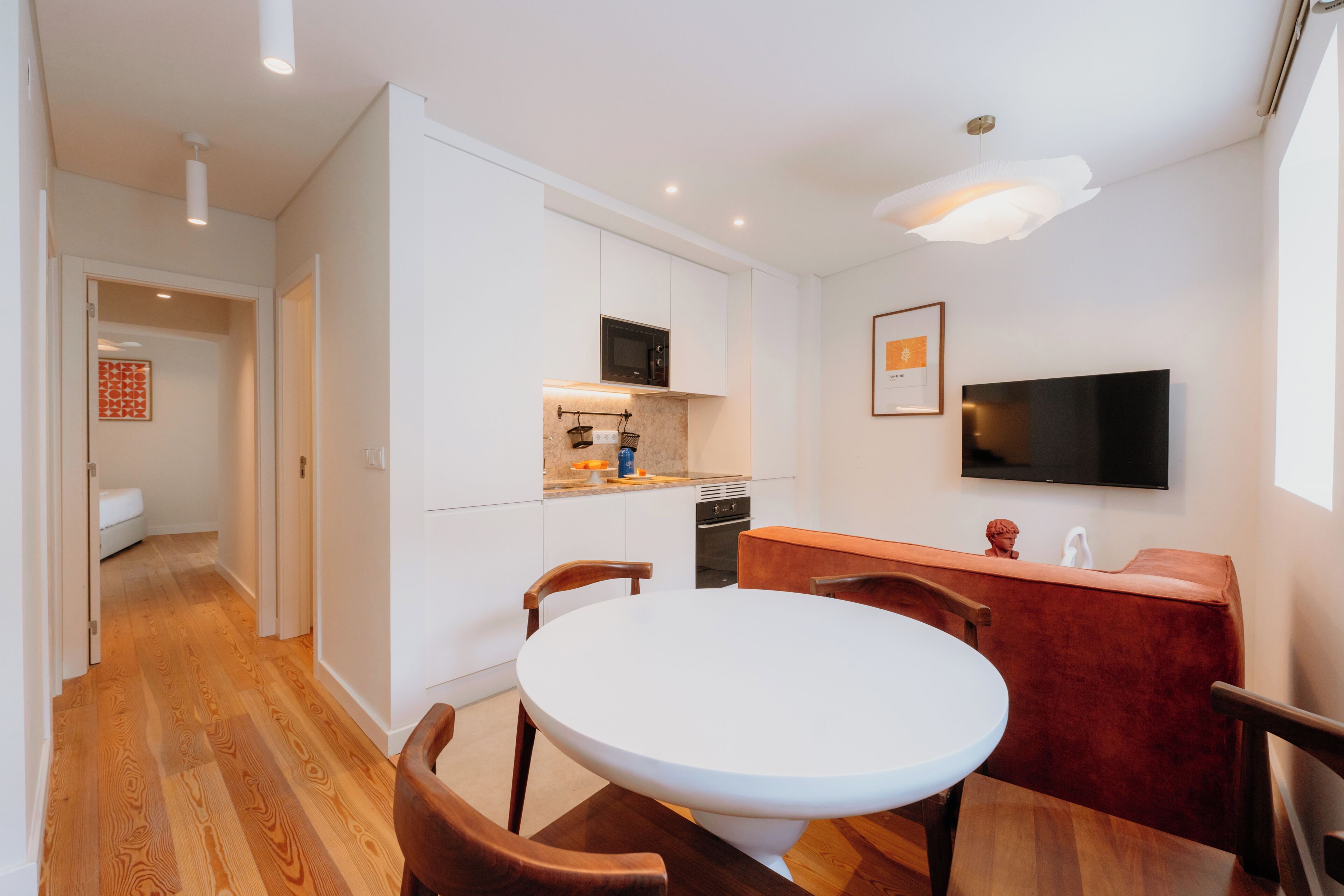 Una cocina moderna con armarios blancos, microondas y horno empotrado. Hay una zona de comedor con una mesa redonda y sillas de madera.