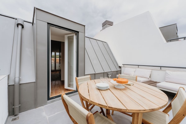 Un área de comedor al aire libre moderna con una mesa redonda de madera, asientos blancos acolchados y vista al cielo.