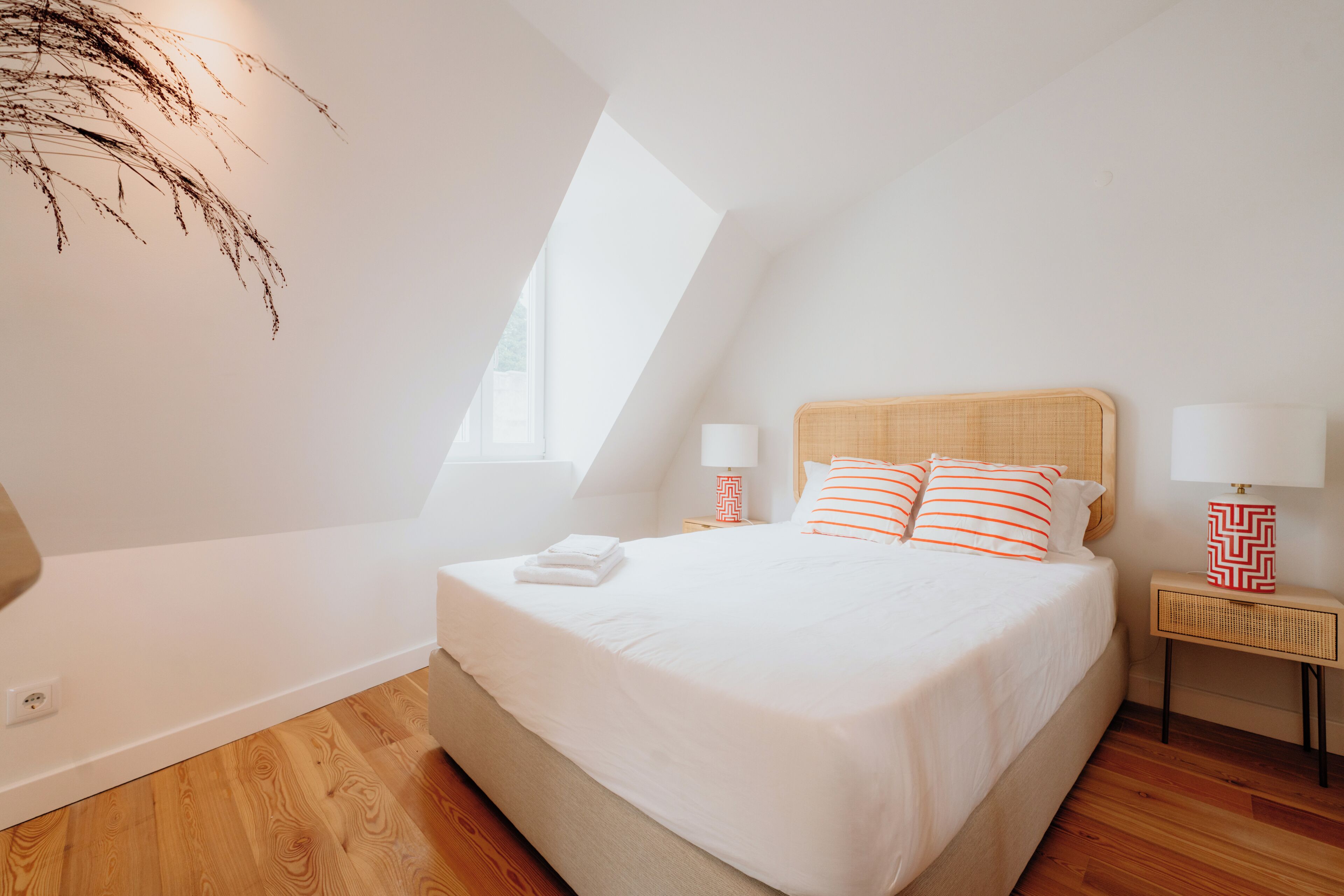 Een slaapkamer met een schuine plafond, een bed met wit beddengoed, twee nachtkastjes met lampen, en een wandlamp.