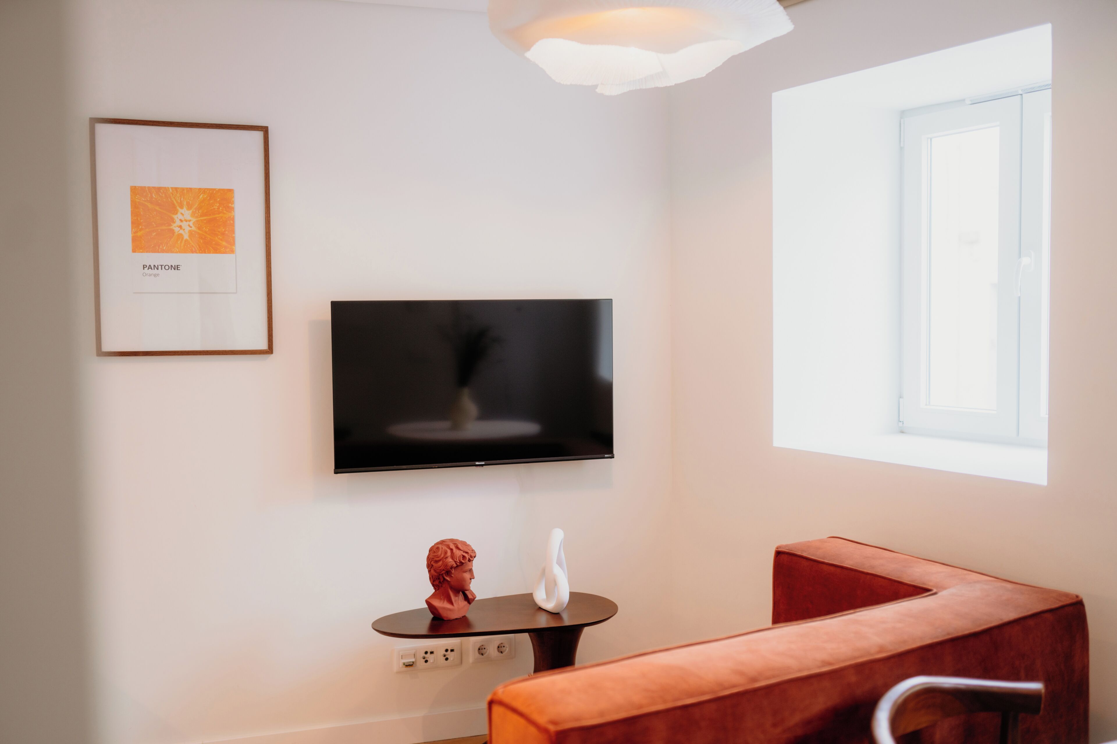 A modern room with a flat-screen TV mounted on the wall, a small table with a bust sculpture, and a cushioned seating area.