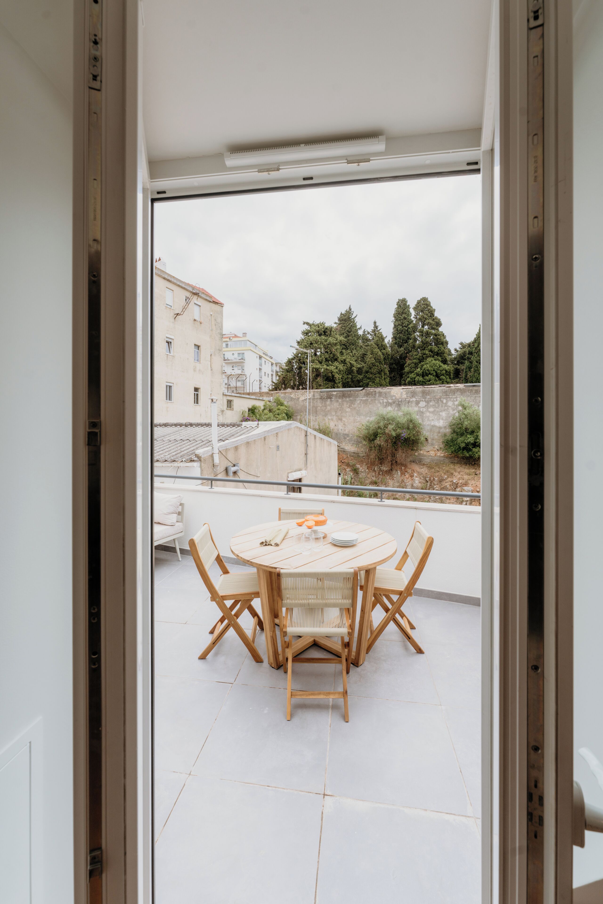 A balcony with a round table and chairs, a view of buildings and trees, and a white tiled floor.