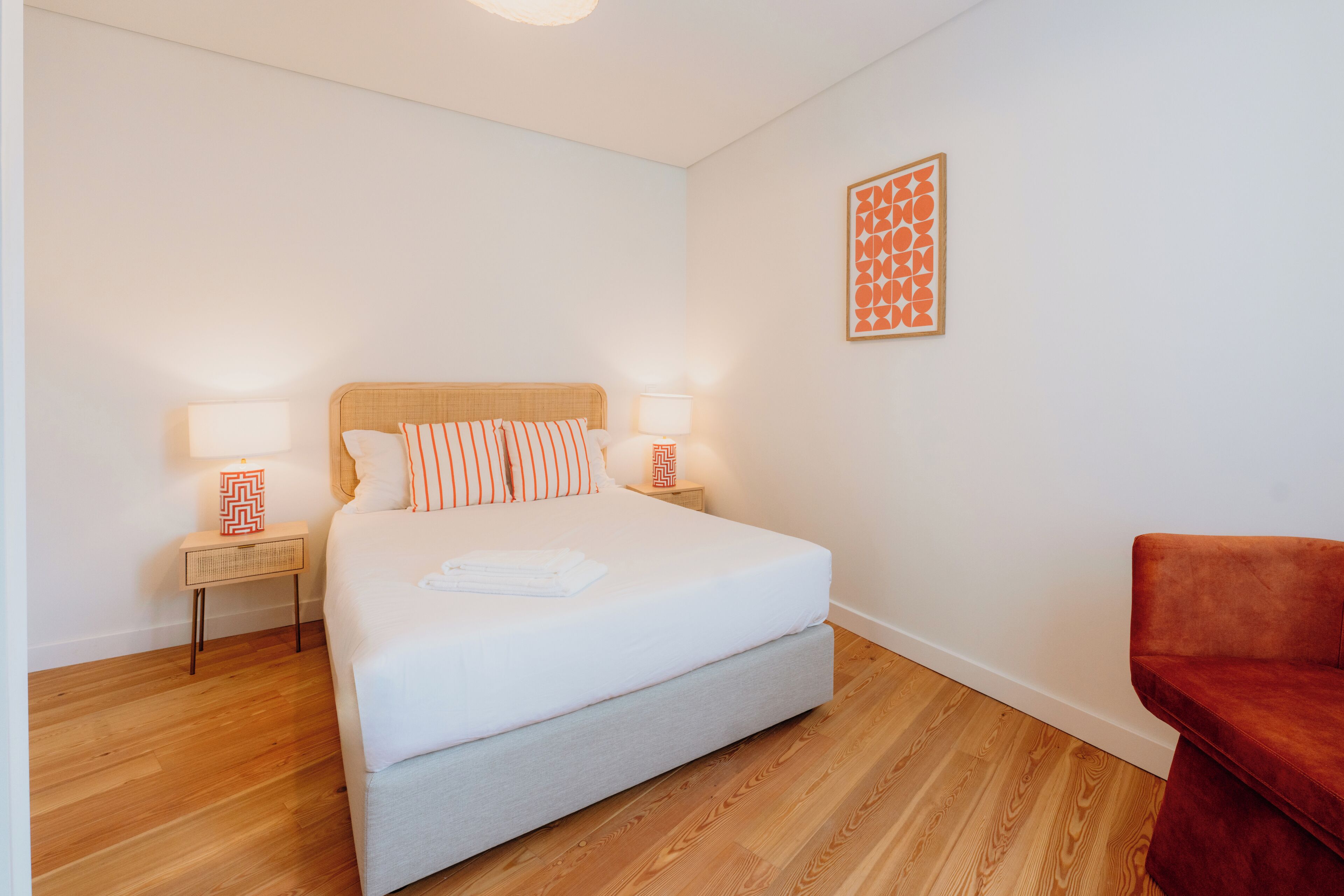 Una camera da letto ordinata con un letto, lampade ai comodini, una poltrona rossa e un'opera d'arte appesa al muro.