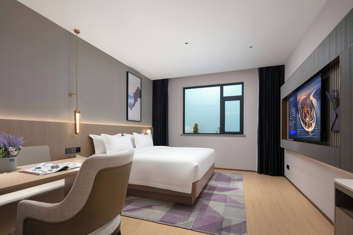 Una habitación de hotel moderna con cama, escritorio, silla, televisor y un ventanal grande.