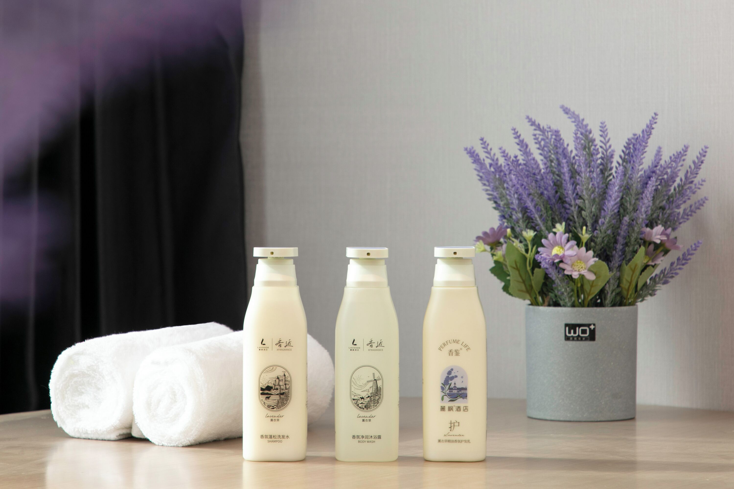 Three bottles of lavender-scented products, a rolled white towel, and a potted lavender plant on a wooden surface.