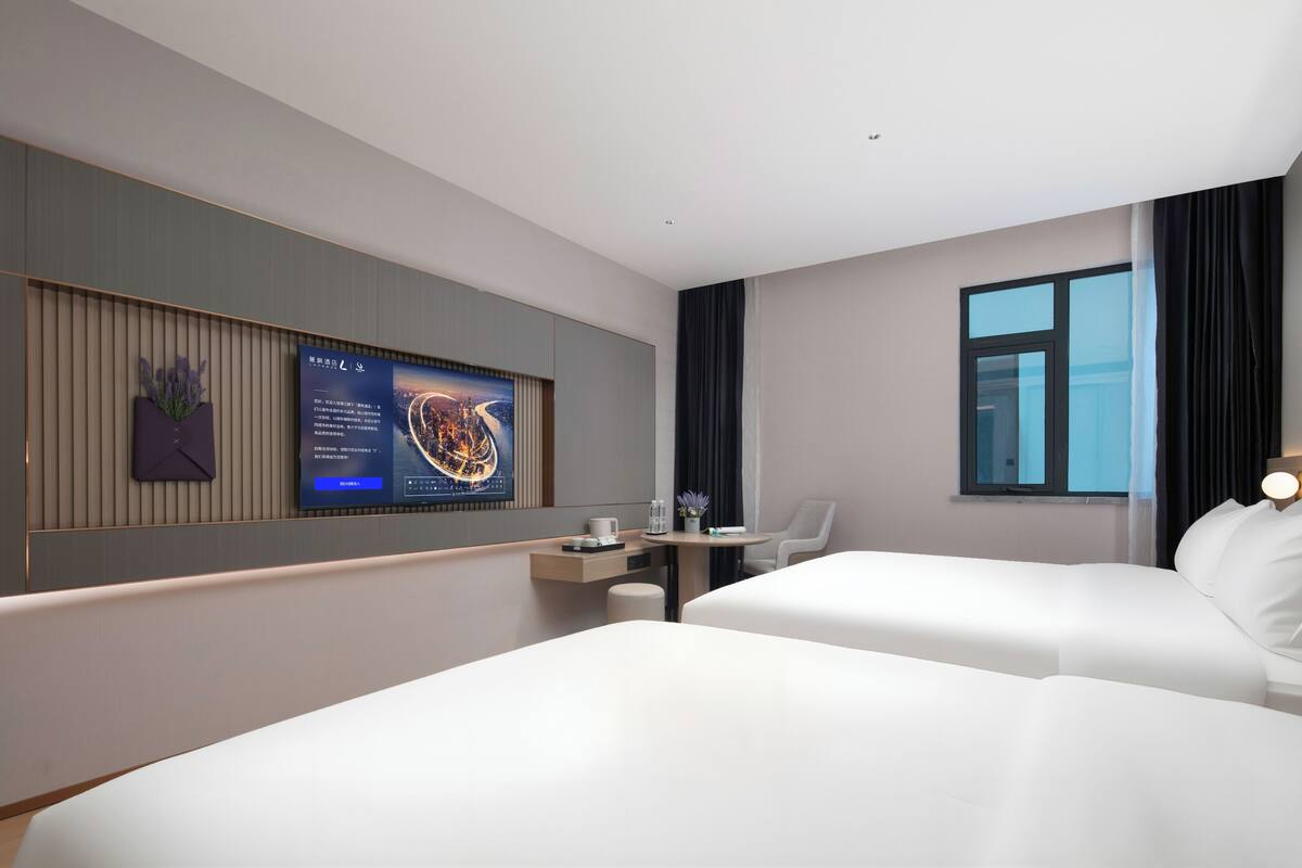 Habitación de hotel con dos camas, un escritorio con silla, televisor montado en la pared y una ventana con cortinas.