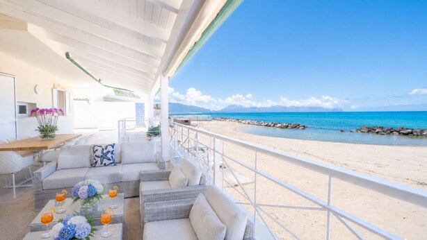 A balcony with a view of the beach, wicker furniture, a table with drinks, and a flower vase. - Amira Beach Villa by LeDhome (Trappeto)