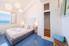 A bedroom with a bed, bedside lamps, a view of the sea, a closet, and a wooden bench with towels. - Amira Beach Villa by LeDhome (Trappeto)