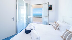 A hotel room with a bed, a glass door leading to a balcony with a view of the sea, and a painting on the wall. - Amira Beach Villa by LeDhome (Trappeto)