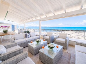 A covered outdoor seating area with wicker furniture, a glass coffee table, and a view of the beach. - Amira Beach Villa by LeDhome (Trappeto)