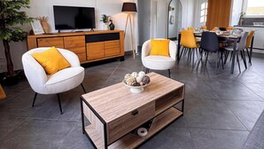 A modern living room with a TV, sofa, coffee table, and dining area with chairs and a table. - Tranquiloue - Maison 8 Pers - 15 min Puy du Fou (Les Herbiers)