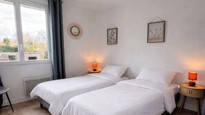 A bedroom with two beds, a window, a wall clock, and a small round table with a lamp. - Tranquiloue - Maison 8 Pers - 15 min Puy du Fou (Les Herbiers)