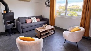 A modern living room with a sofa, two armchairs, a coffee table, a fireplace, a clock, and a large window with curtains. - Tranquiloue - Maison 8 Pers - 15 min Puy du Fou (Les Herbiers)