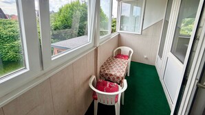 Una habitación con vistas a zonas verdes y edificios, una silla blanca con mantel de flores, y una alfombra verde.