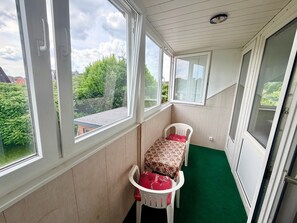 A room with a view of greenery and buildings, a white chair with a floral tablecloth, and a green carpet.