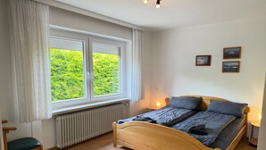 Makuuhuoneessa on puinen sänky, tuoli, ikkuna verhoilla, patteri ja seinään kiinnitetyt valaisimet.