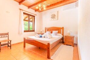 A bedroom with a wooden bed, a nightstand, a bench, a window with curtains, and a ceiling fan. - Villa Son Segi by Slow Villas (Sant Llorenç Des Cardassar)