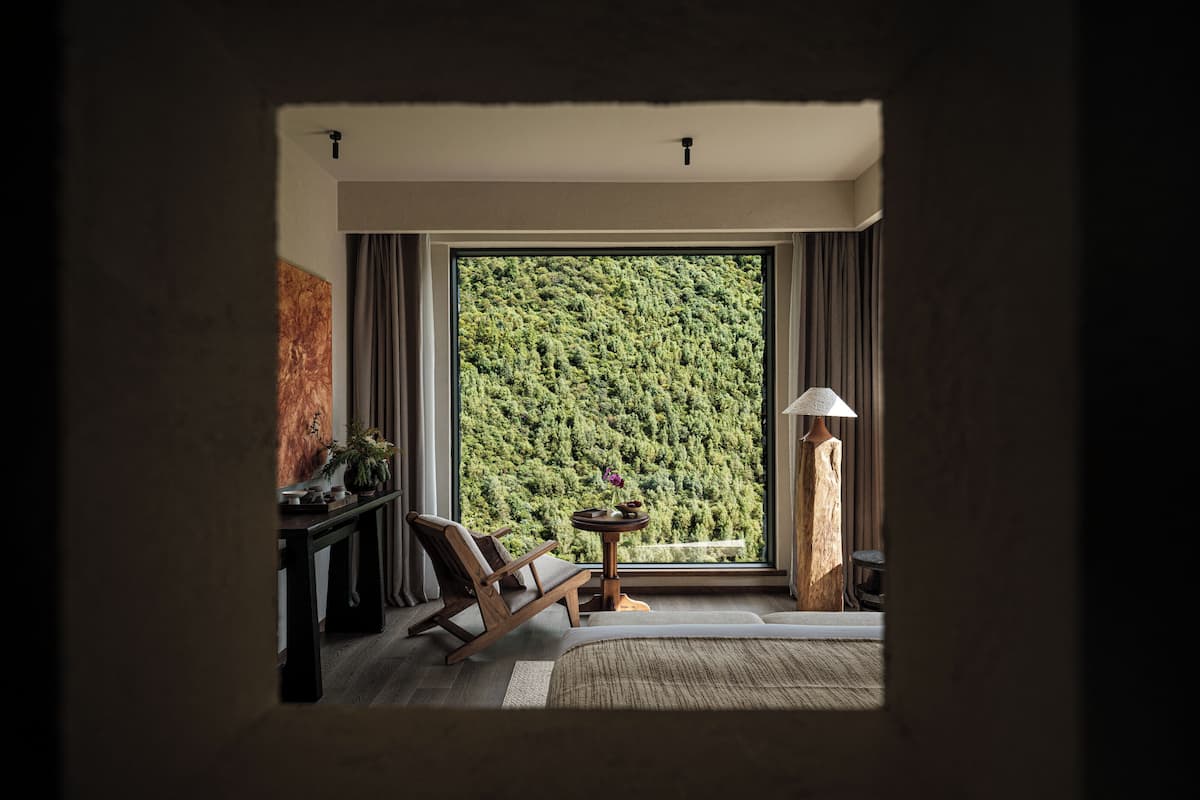 Ein Zimmer mit großem Fenster, einem Holzstuhl, einem kleinen Tisch mit Lampe und einer Pflanze.