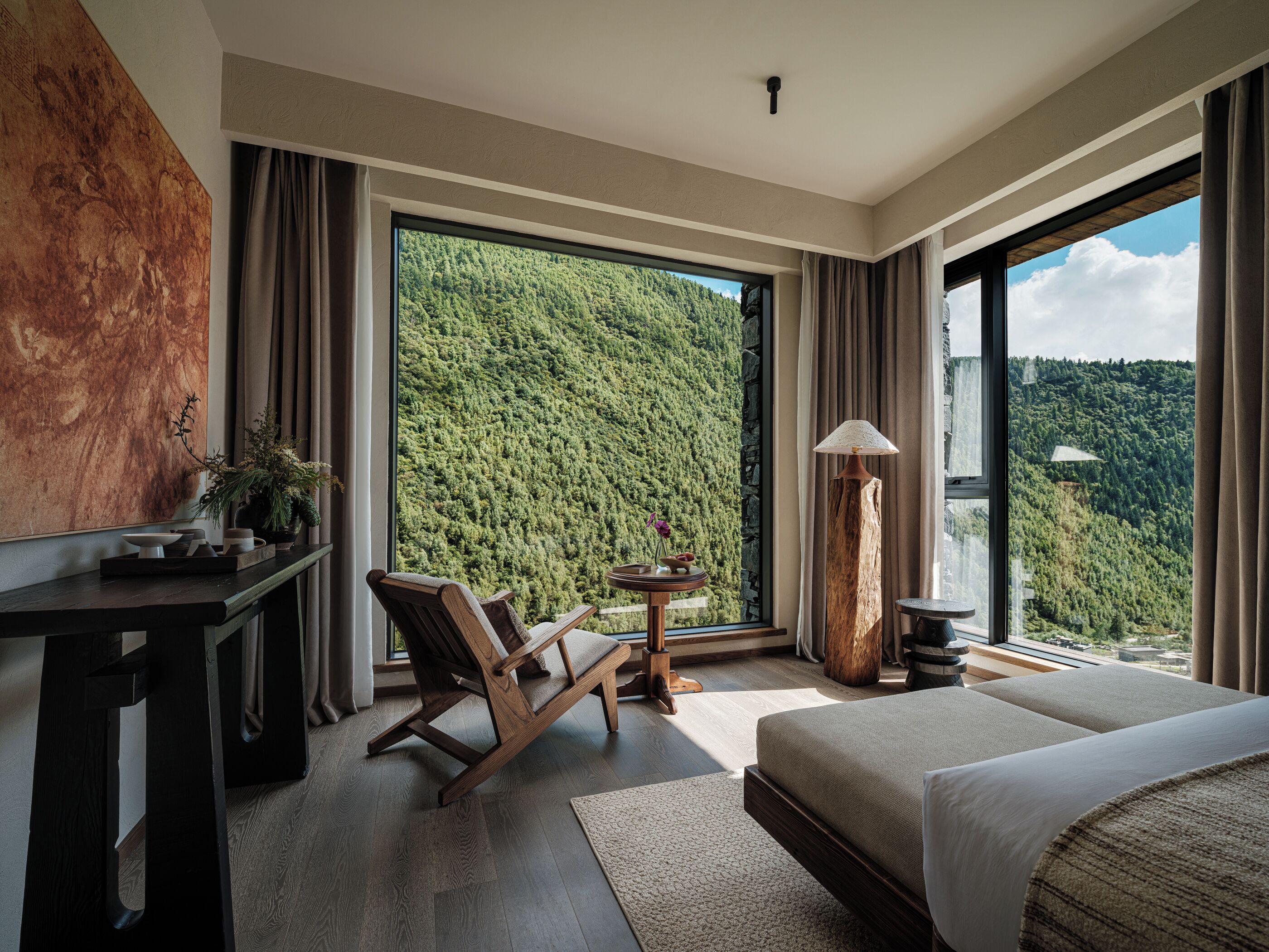 A modern hotel room with a large bed, a desk, a chair, a small table with a lamp, and a view of a forested hillside.