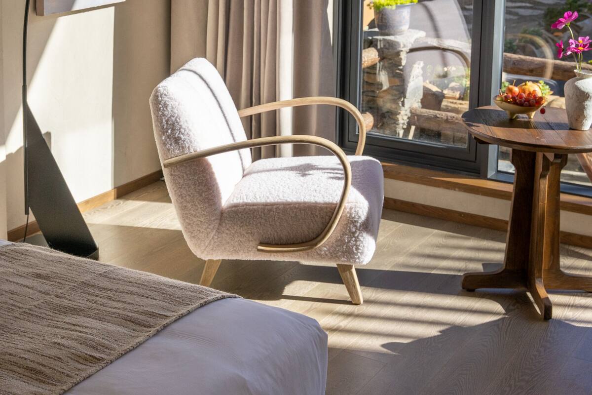Ein modernes Hotelzimmer mit einem Bett, einem Sessel, einem Tisch mit Obstschale und einem Blumentopf. Durch ein großes Fenster ist viel Grünlandschaft zu sehen.