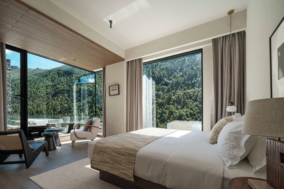 Ein modernes Schlafzimmer mit einem großen Bett, einem Schreibtisch und einem Sessel, mit Blick auf eine herrliche Berglandschaft durch große Fenster.