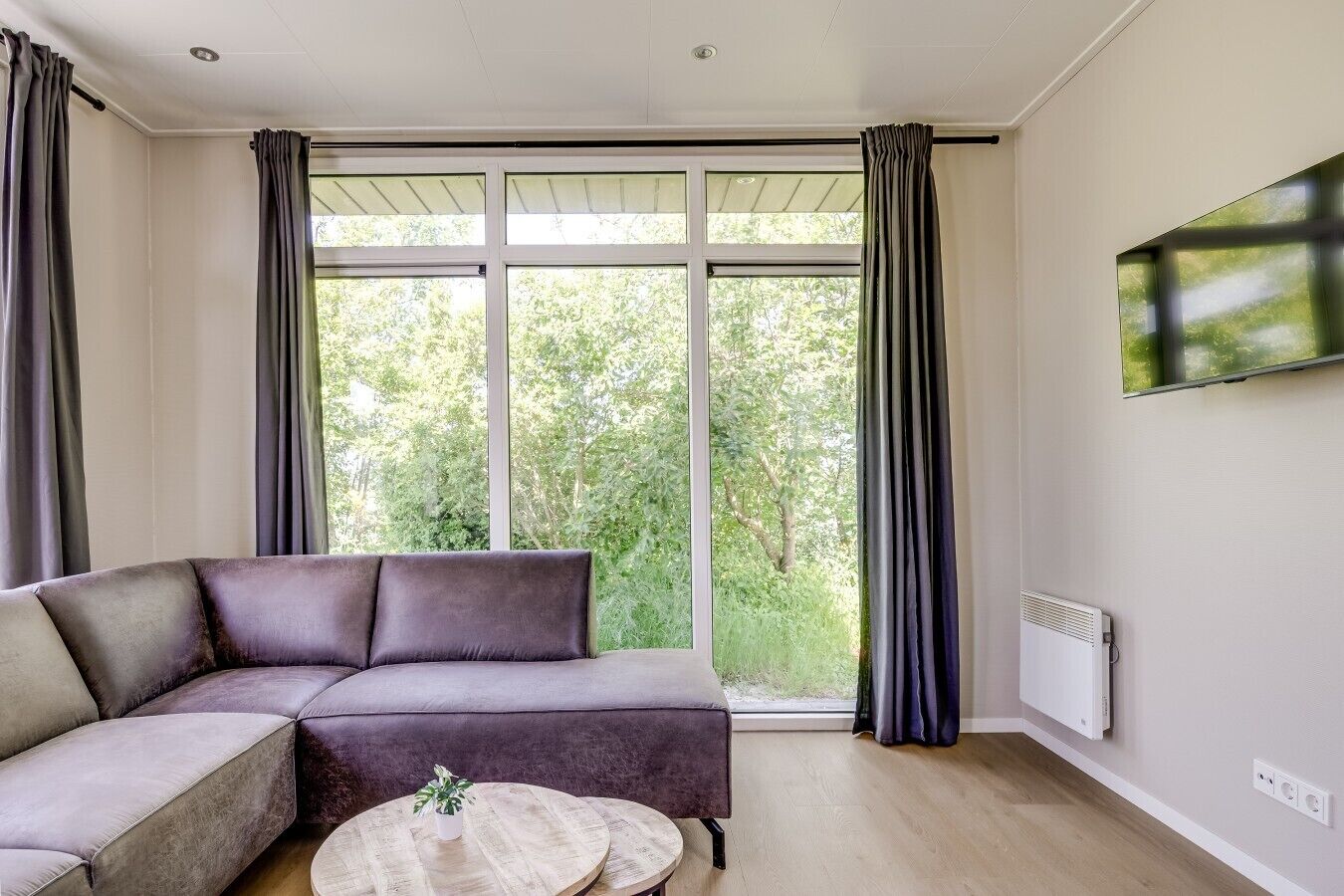 Ein modernes Wohnzimmer mit Ecksofa, rundem Couchtisch und großen Fenstern mit Vorhängen.
