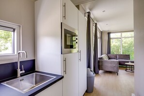 A modern kitchen with a sink, cabinets, and a window. There is a living area with a sofa and a table, and a window with curtains.