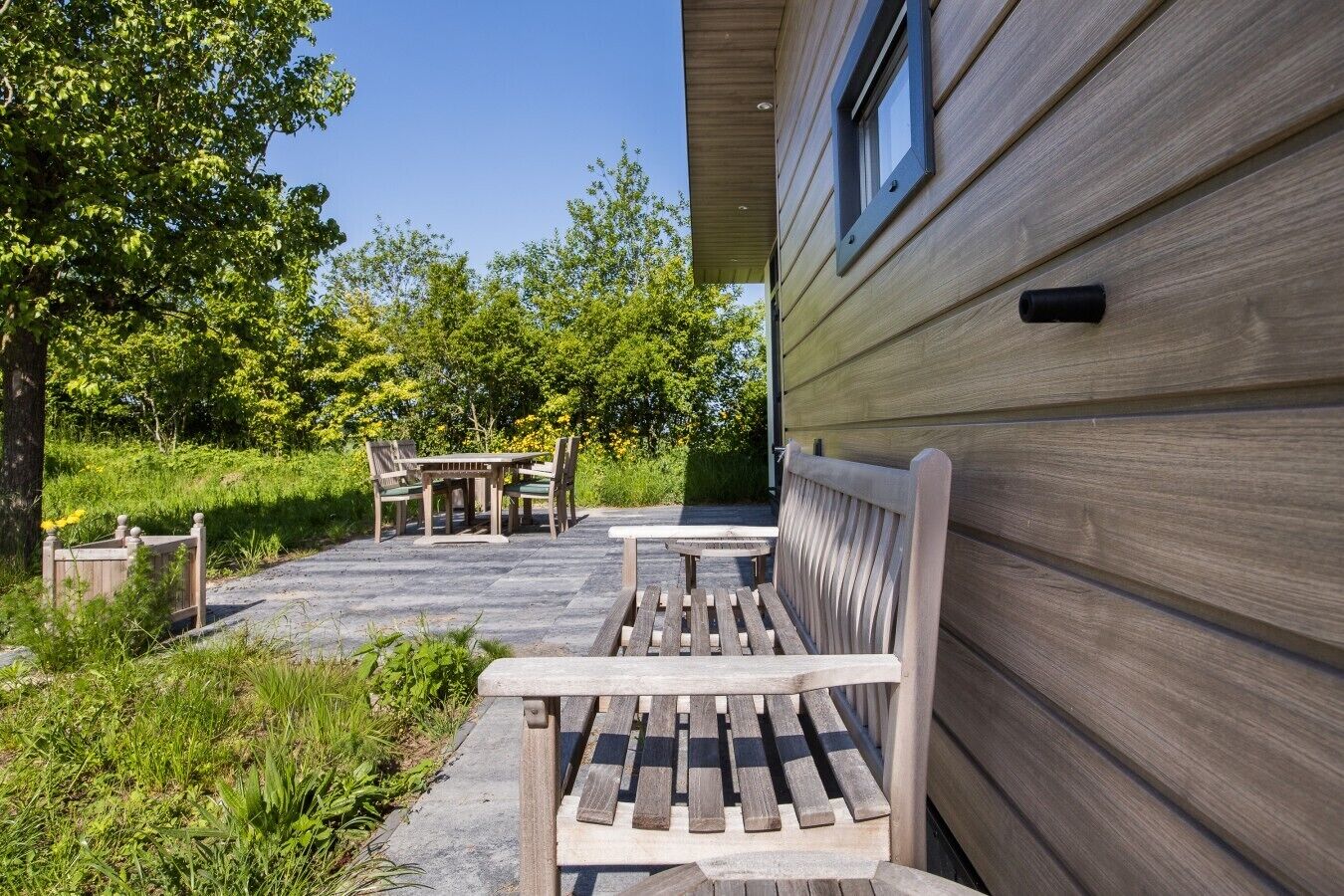Une terrasse en bois avec un banc et une table, entourée de verdure et d’arbres.