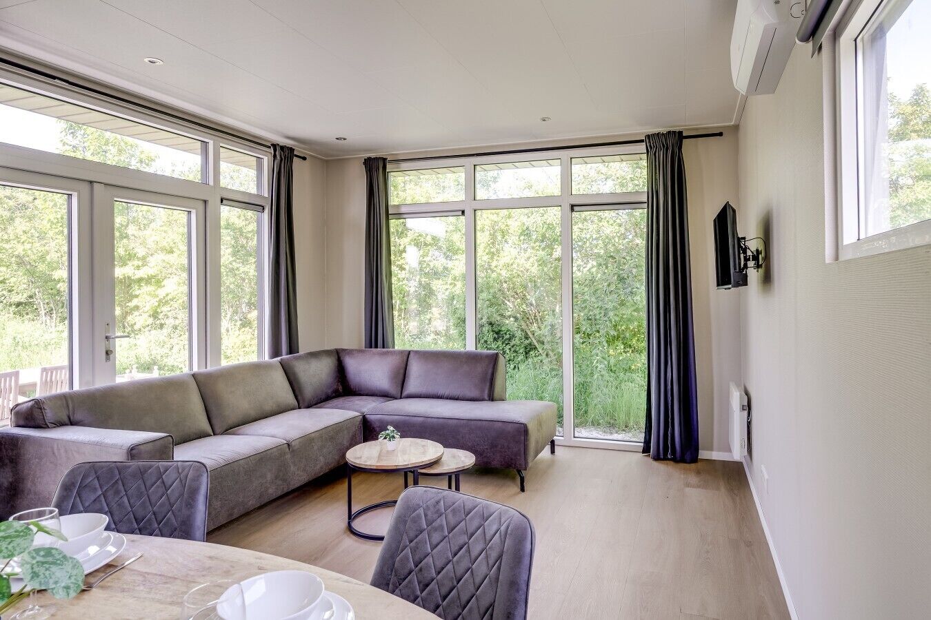 Een moderne woonkamer met een hoekbank, een ronde salontafel en een eettafel gedekt voor twee personen. Grote ramen bieden uitzicht op groen buiten.