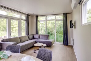 A modern living room with a sectional sofa, a round coffee table, and a dining table set for two. Large windows provide a view of greenery outside.