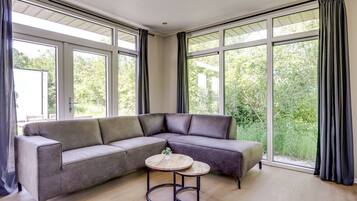 Een moderne woonkamer met een hoekbank, een ronde salontafel en grote ramen voorzien van gordijnen.